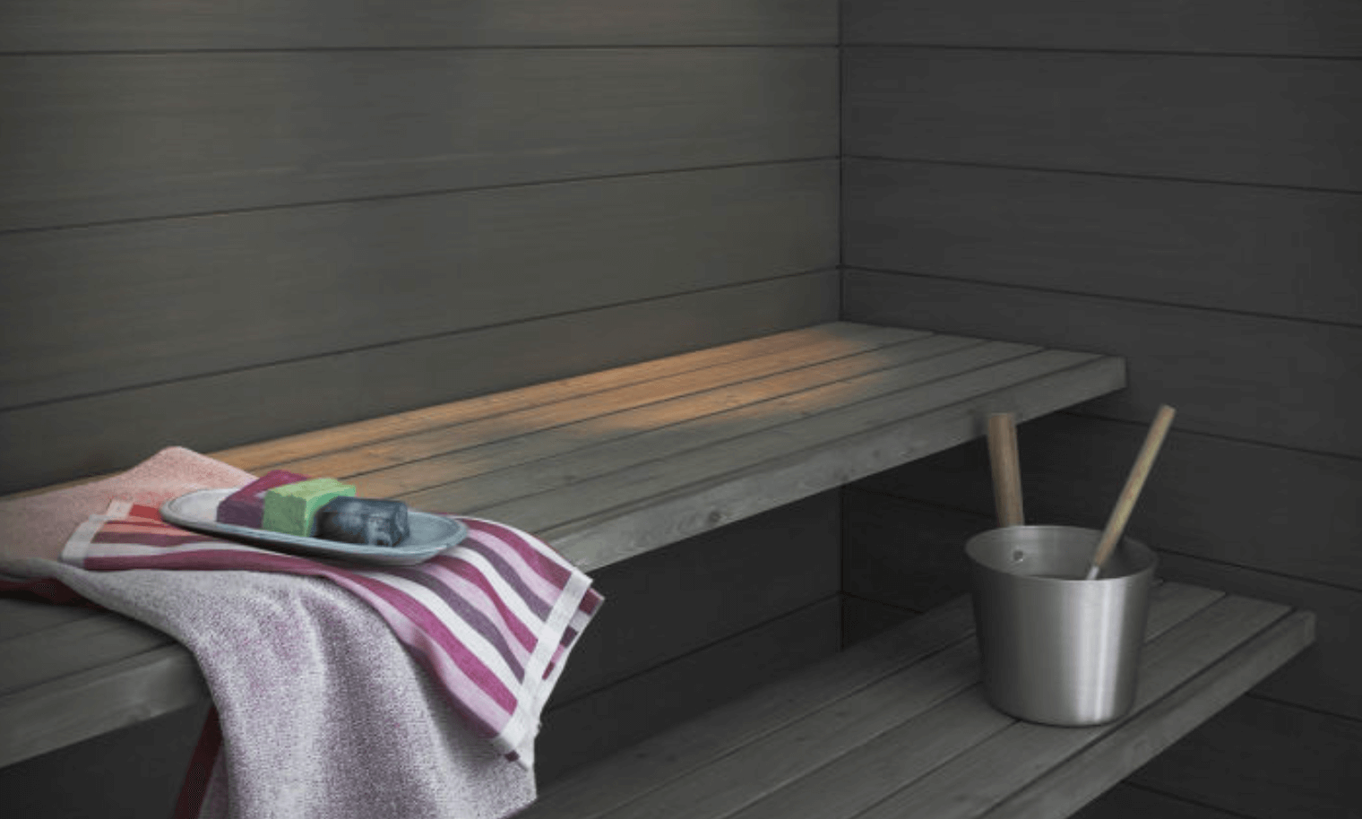 Grå bastulave med rosa-vit randig handduk, tvålar på bricka och metallhink med träskopa.