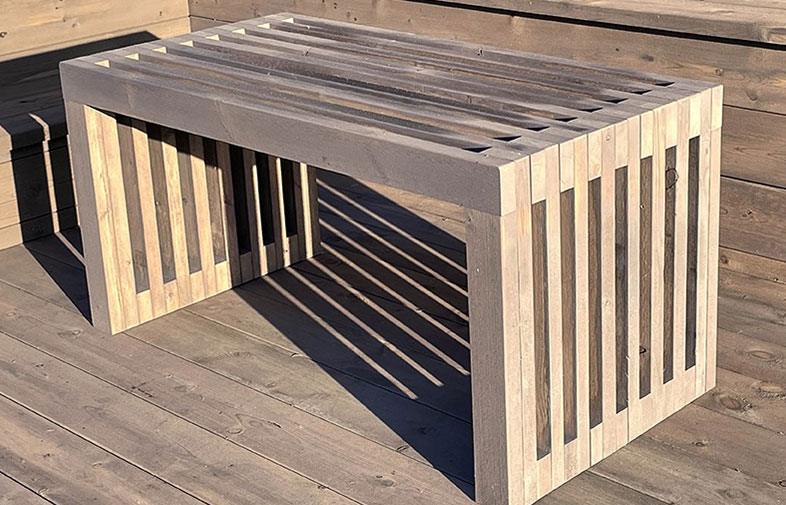 Gråvit slitsad träbänk på träterrass i sol, skuggspeglar från bänkens ribbor.