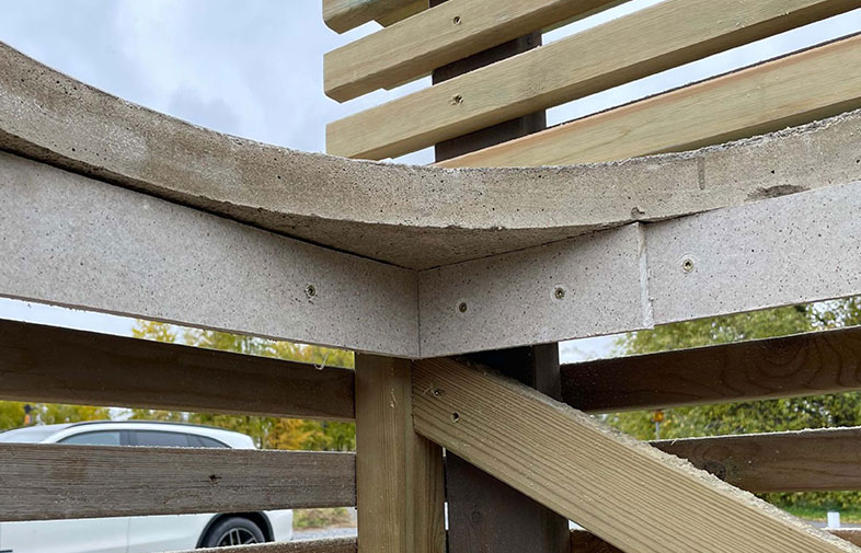 Detalj av utomhusbänk med grå betongsits och ljusbruna trälameller mot grönska och vit bil