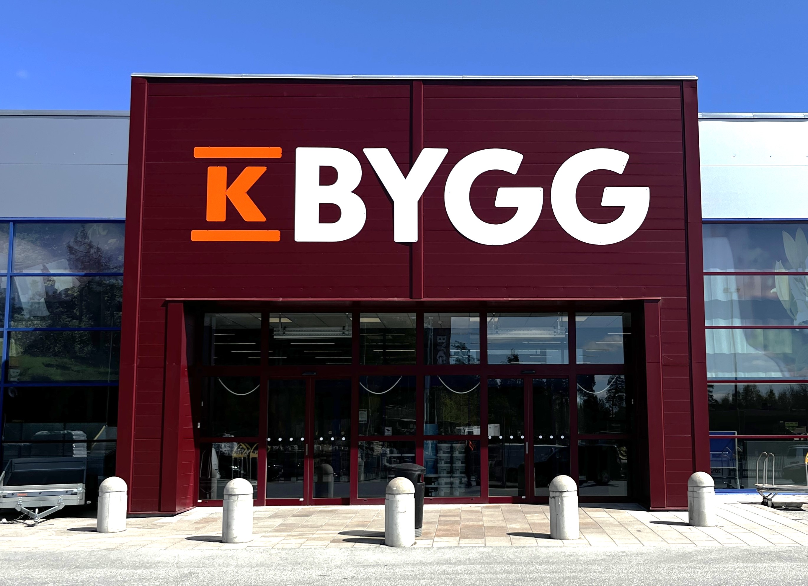 Tre personer i svart/orange arbetskläder står framför en byggfirma-skylt “K BYGG” utomhus.