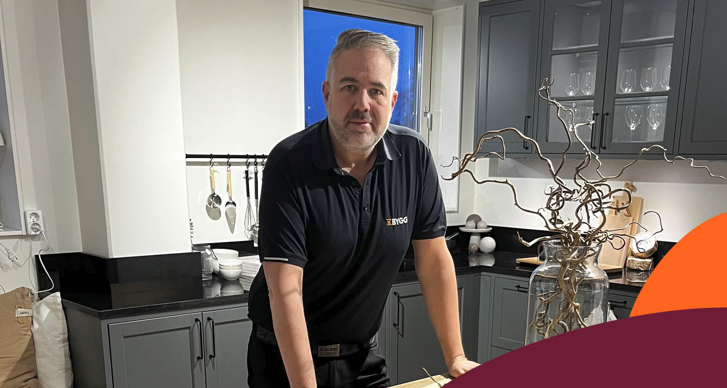 Man i svart pikétröja vid svart bänkskiva i grått kök med glasluckor och vas med kvistar