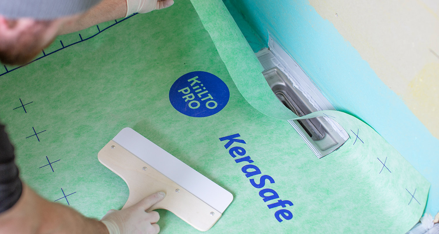 En hand i handske slätar ut grön KeraSafe-tätmembran med spackelspade runt golvavlopp.