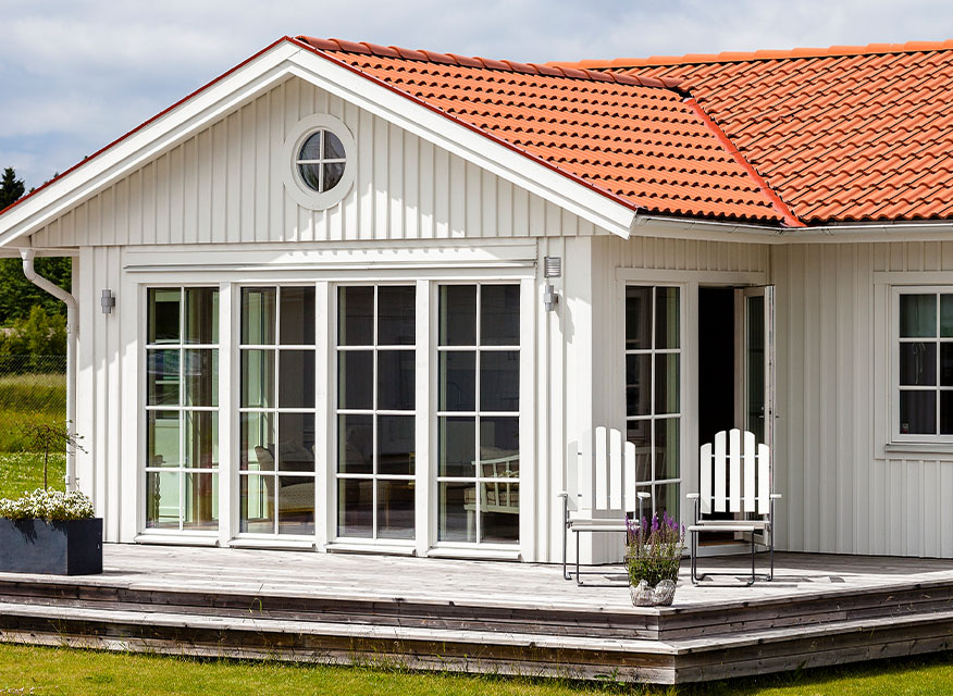 Vit trävilla med rött tegeltak, stora glasdörrar mot altan med vita stolar och blommor.