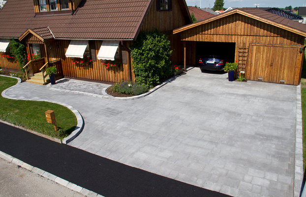 Brunt trähus med vita markiser, röda blommor i fönster, grå uppfart och öppet garage med svart bil.
