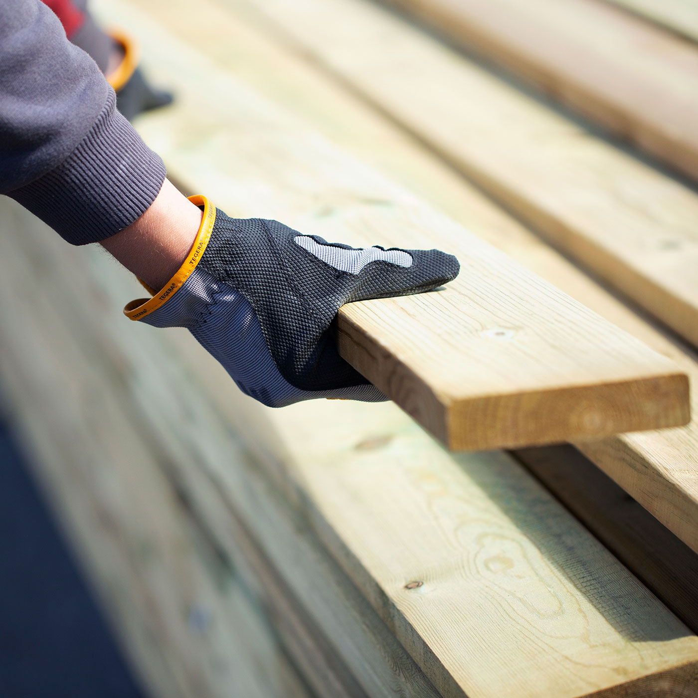 Svartgrå arbetsvante med gula detaljer håller ljus träplanka över stapel träbalkar