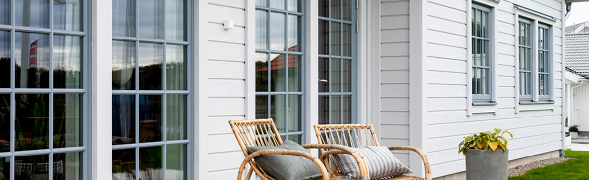 Vit träfasad med stora grå fönster, två rottingstolar med kuddar och kruka på grön gräsmatta