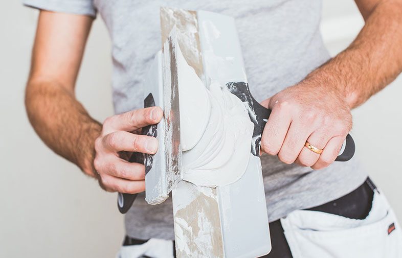 Närbild på byggarbetare i grå t-shirt som med spackelspade applicerar vit spackel på bräda