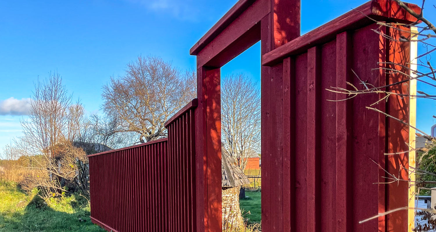Rött trästaket med öppen grind mot grön trädgård, nakna träd och klarblå himmel.