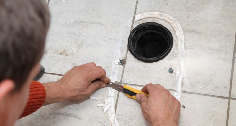 Två händer skär bort vit fogmassa med gul kniv runt en mörk, rund golvbrunn på ljusa kakelplattor.