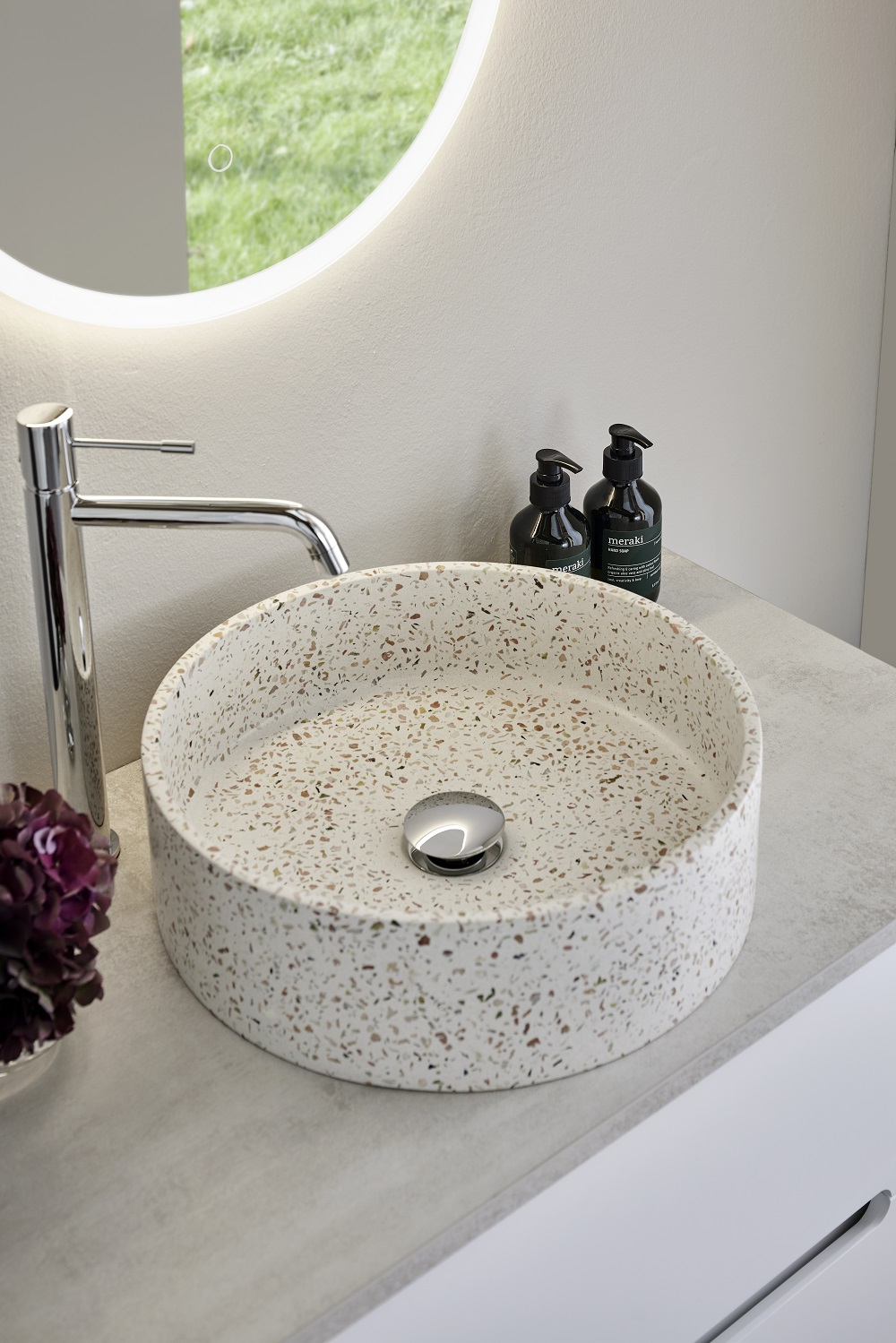 Rund vit terrazzohandfat med färgprickar på grå bänk, kromkran, svarta pumpflaskor och belyst spegel