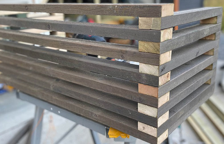 Mörkbrun trälåda med horisontella slatar på metallstativ i verkstad.