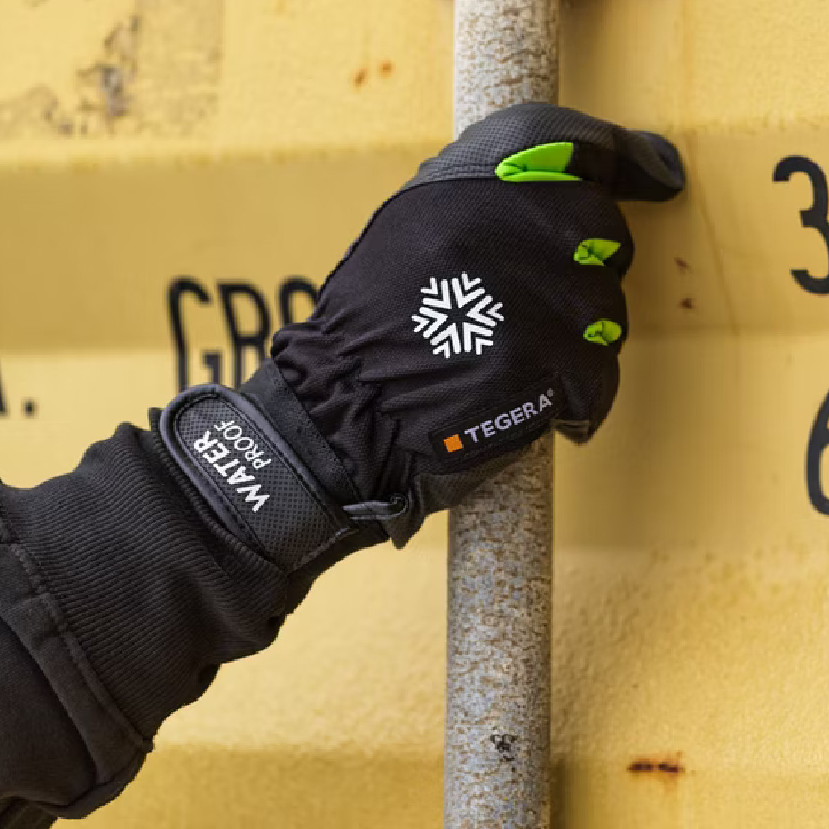 Hand i svart och grön ’TEGERA’-handske greppar rostig stång framför gul container.