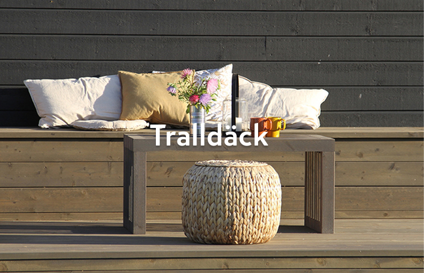 Solbelyst trädäck med bänk, vita och gula kuddar, bukett i plåtvas, rottingpall och muggar