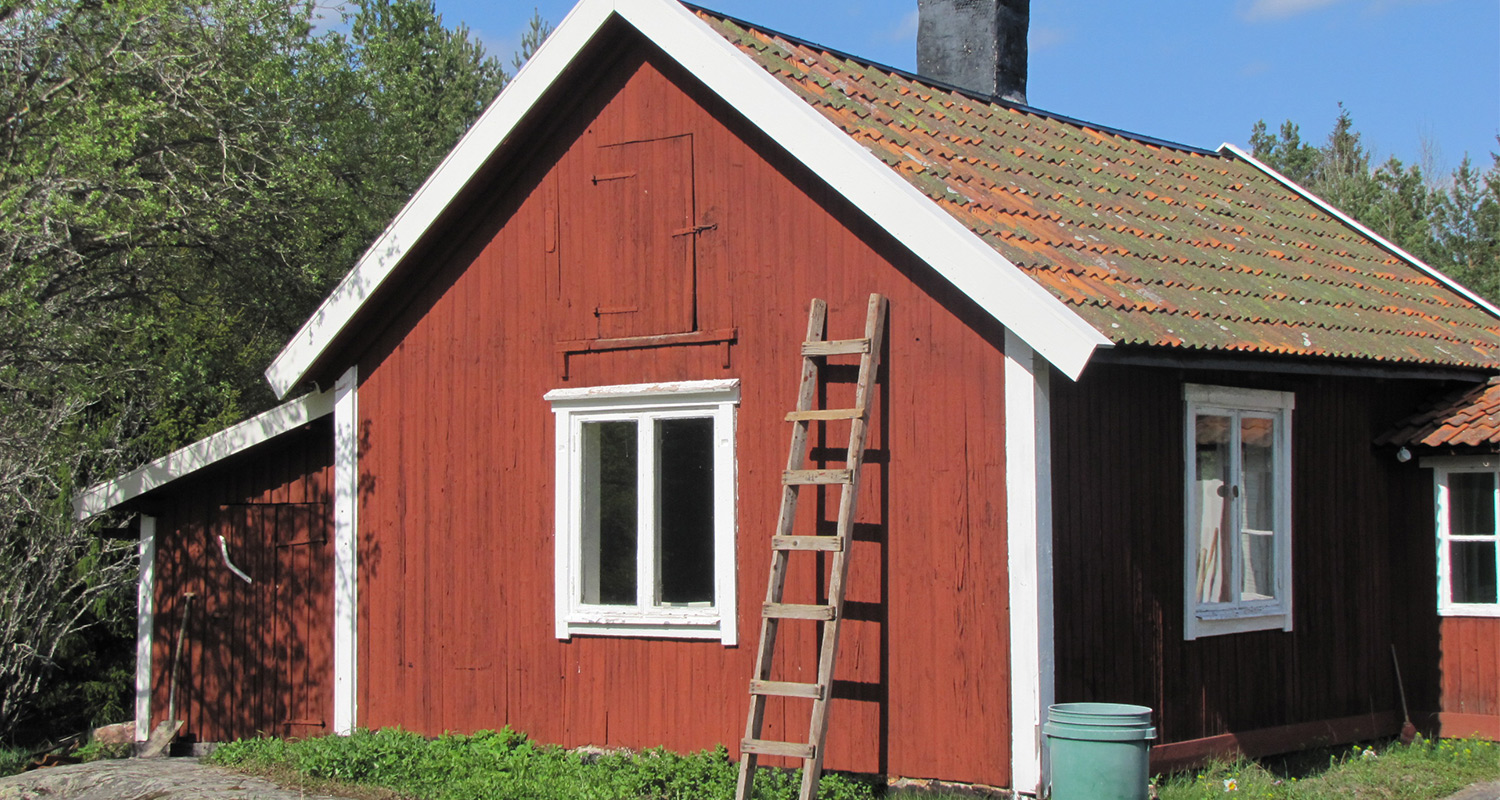 Röd trästuga med vita fönsterramar, mossigt tegeltak och trästege lutad mot väggen.