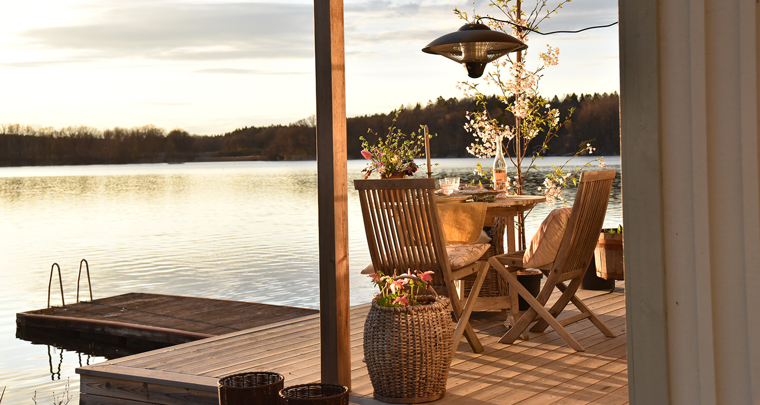 Solbelyst träaltan vid sjö med dukat bord, blombuketter, stolar och stilla vatten.