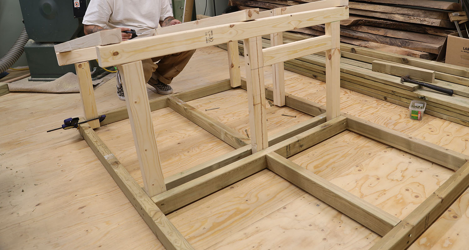 Ljust träram under montering på plywoodgolv i verkstad, del av person i vit tröja syns