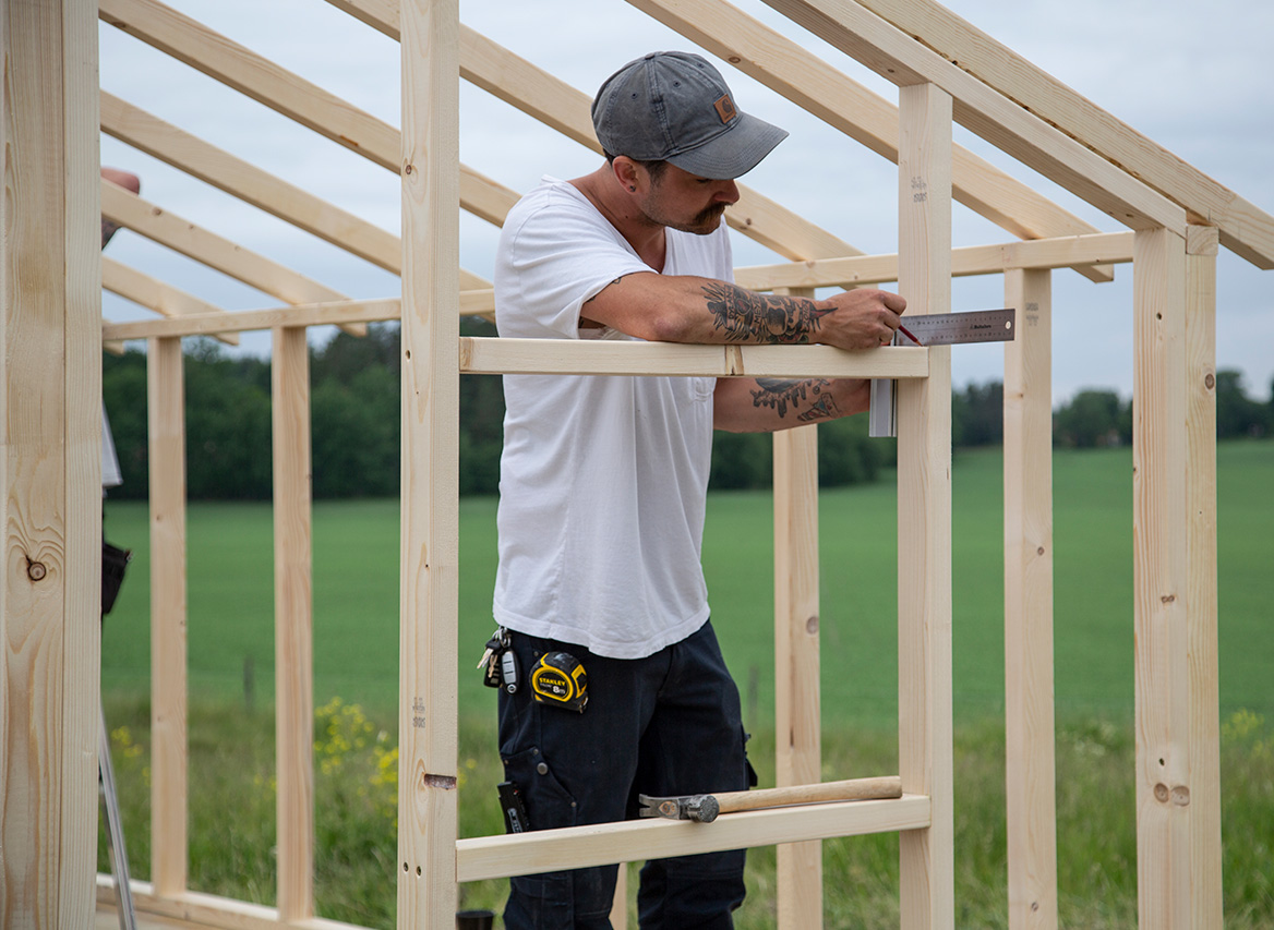 Man i vit t-shirt och grå keps mäter träreglar på ramverk i grön sommaräng