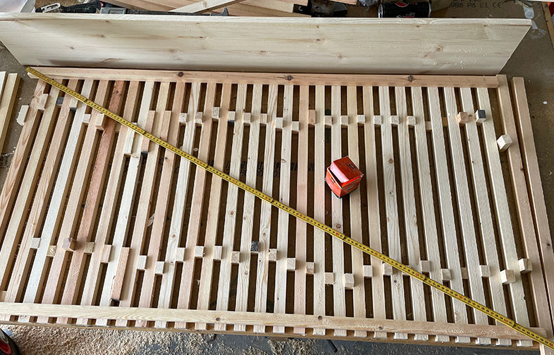 Ljus träram med parallella ribbor, gul tumstock diagonalt och rött måttband ovanpå i verkstad