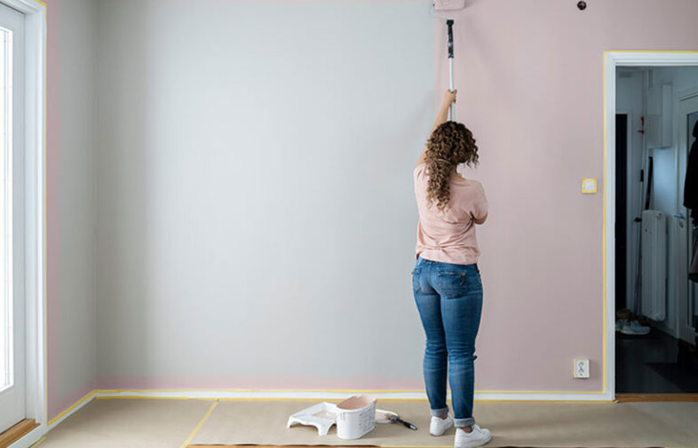 Kvinna i jeans och rosa tröja målar med roller en ljusgrå till rosa vägg i bostadsrum.