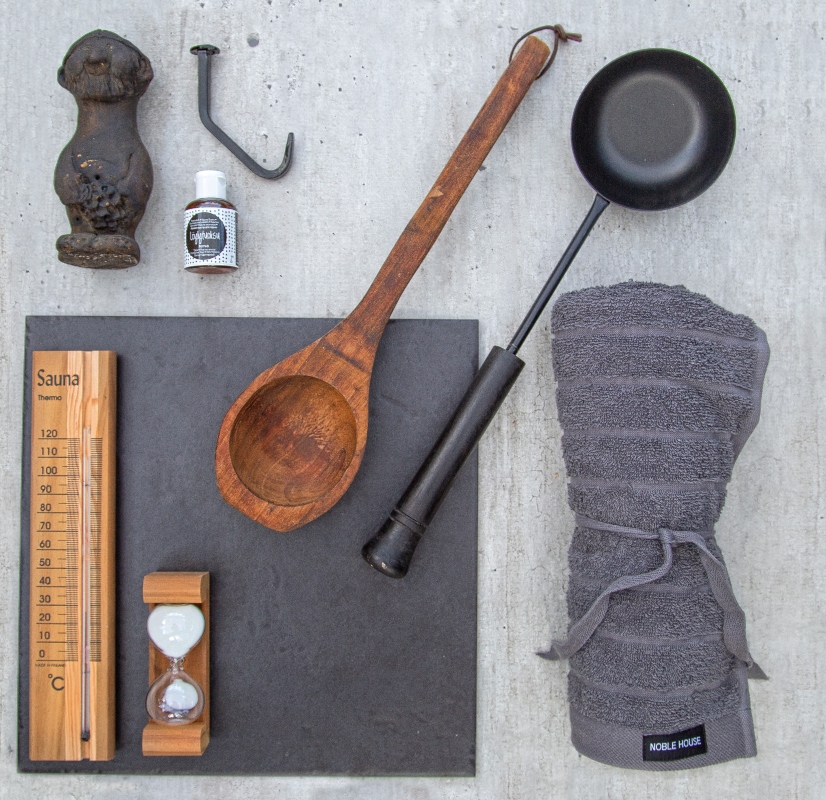 Bastuattiraljer: oljeflaska, gjutjärnsfigur, träskopa, svart ösa, termometer, timglas och grå handduk