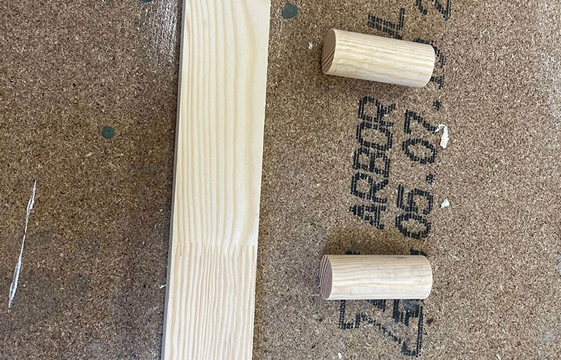 Ljust träregel och två träpluggar på brunt spånskiva med stämplat text.