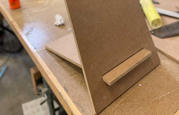 Närbild på brunt MDF-ställ på träbänk i verkstad med gul överstrykningspenna och verktyg.