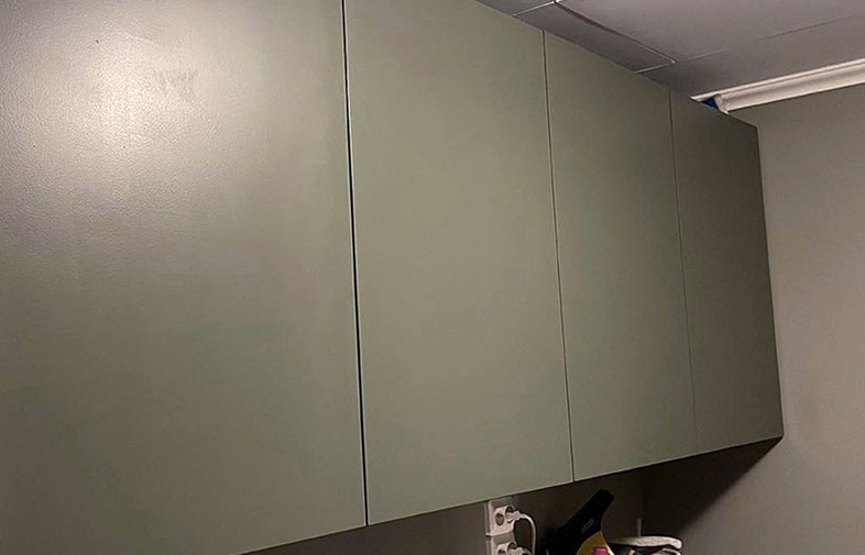 Fyra väggmonterade olivgröna skåpluckor i tvättstuga, med eluttag och flaskor under