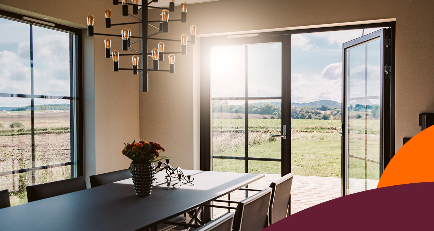 Modern matplats med svart bord, vas med blommor, takkrona och öppna glasdörrar mot grön åker.