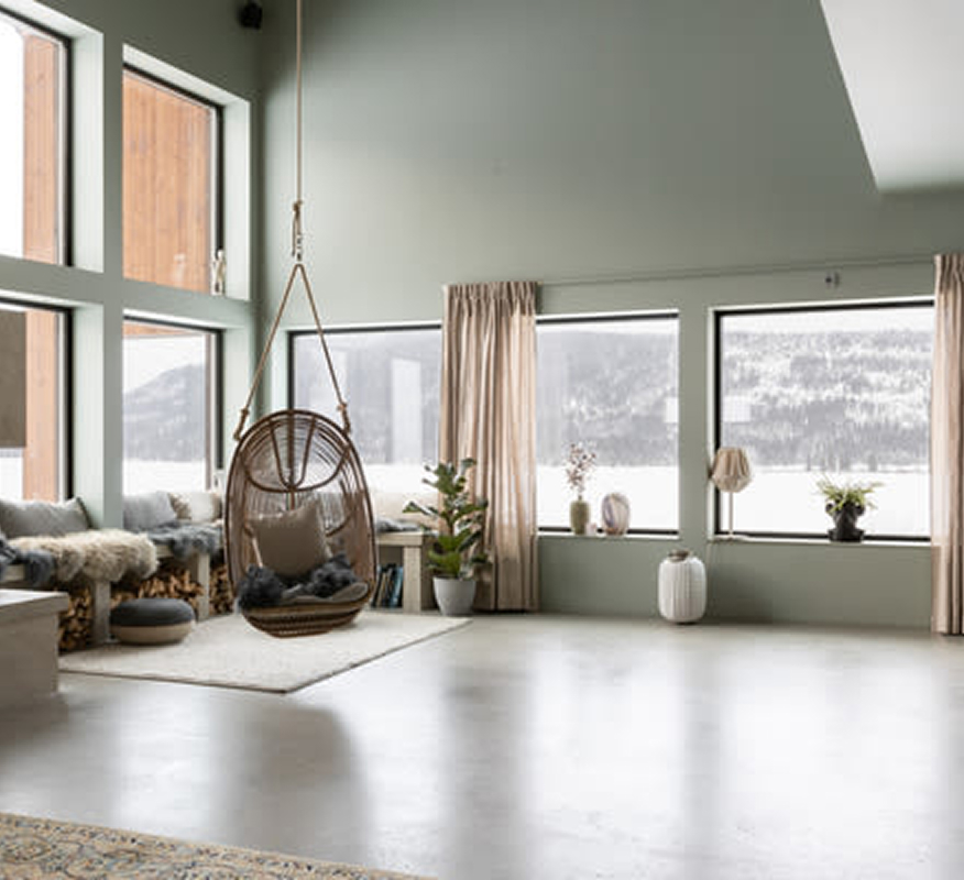 Ljust rum med gröna väggar, stora fönster mot snölandskap, rottinghängstol och växter.