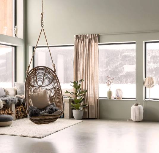 Rumsbild med rottinghängstol, ljusa nyanser och stora fönster mot snölandskap.