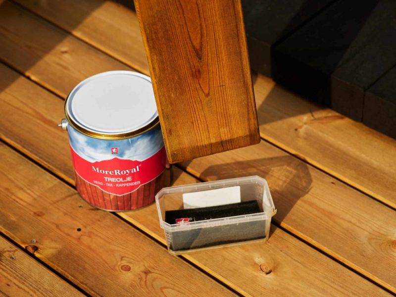 Rödblå MoreRoyal träoljeburk med penselsvamp och oljad bräda på solbelagd trätrall