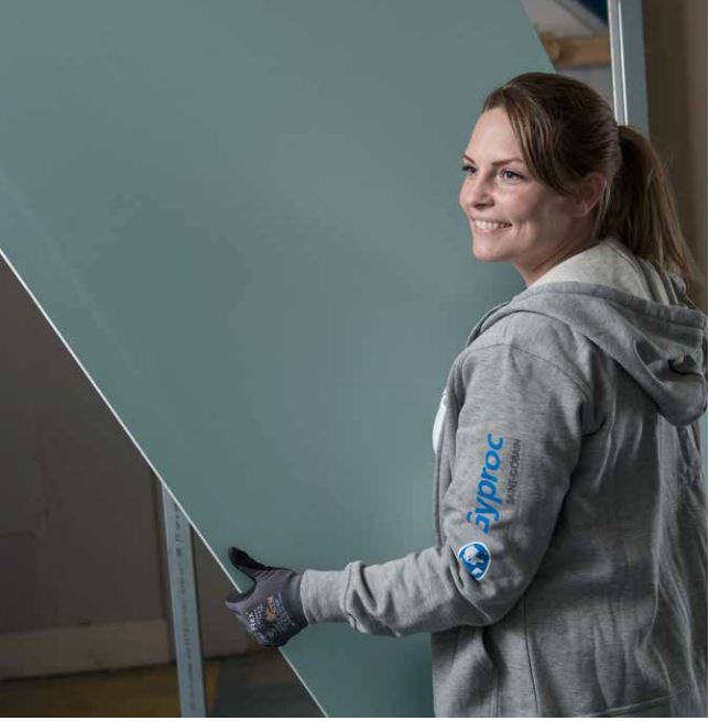 Arbetare i grå Gyproc-hoodie bär stor grön gipsskiva i byggmiljö