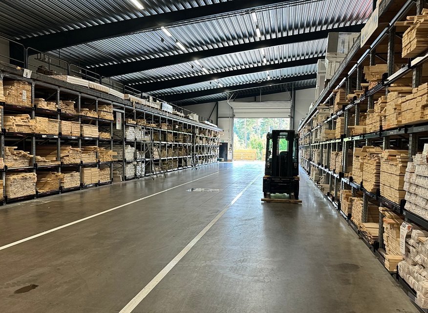 Lång lagerhall med metallhyllor fyllda av träbrädor och en gaffeltruck i gången