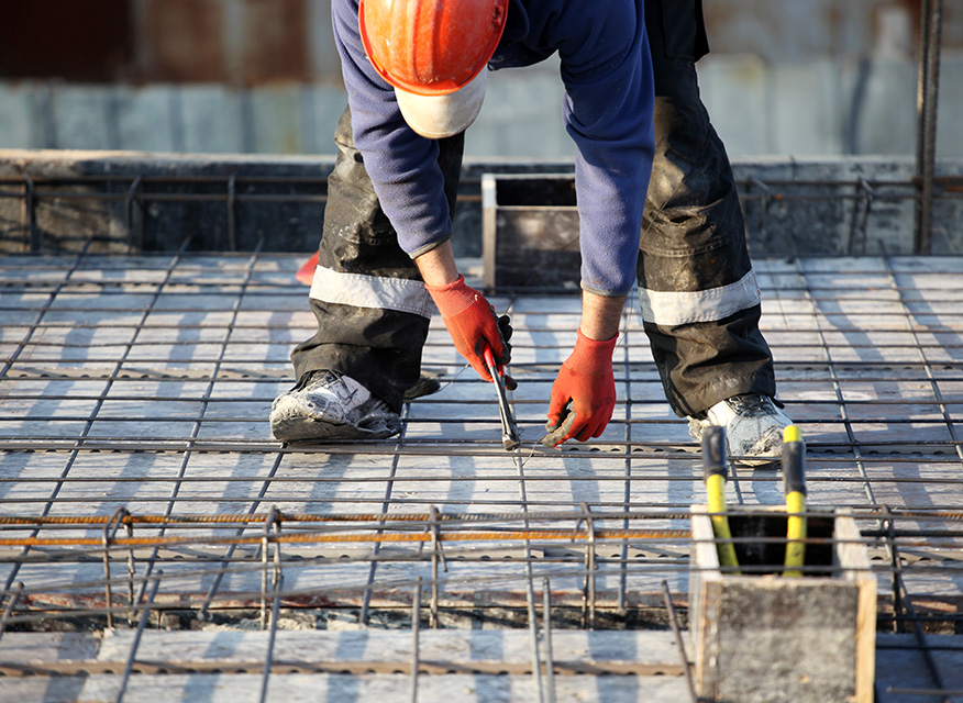 Byggarbetare i orange hjälm och röda handskar binder armeringsjärn på betonggolv.