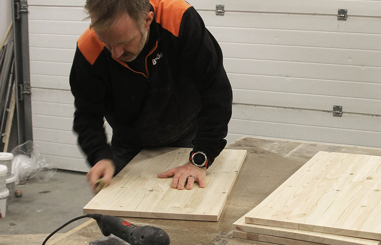 Arbetare i svart-orange arbetsklädsel slipar ljus träskiva på ett verkstadsbord.