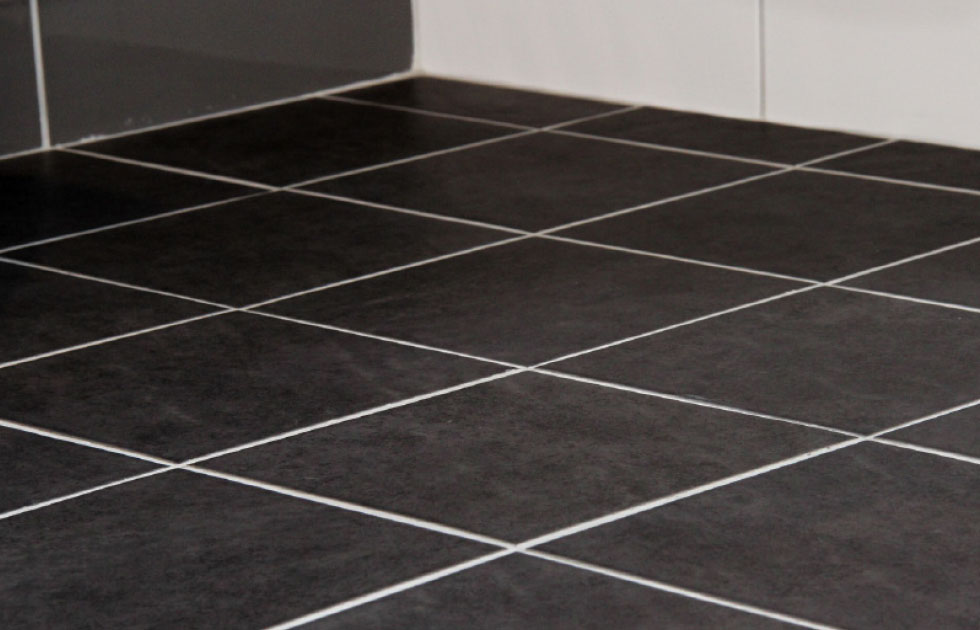 Mörka kvadratiska golvplattor i svartgrå ton med vita fogar mot vit vägg.