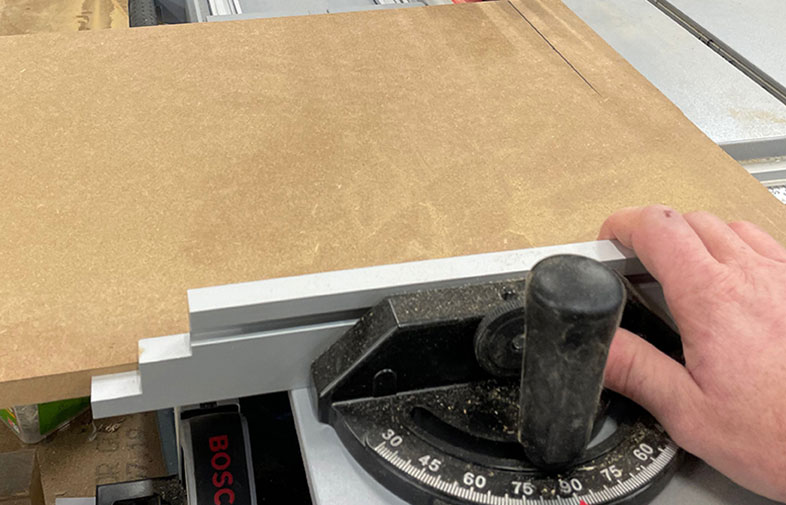 Hand håller beige MDF-skiva mot svart-silver gersågsguide på bordssåg.