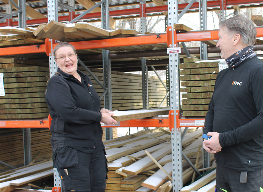 Två personer i svarta kläder håller träplanka framför orange metallhyllor fyllda med ljusa brädor