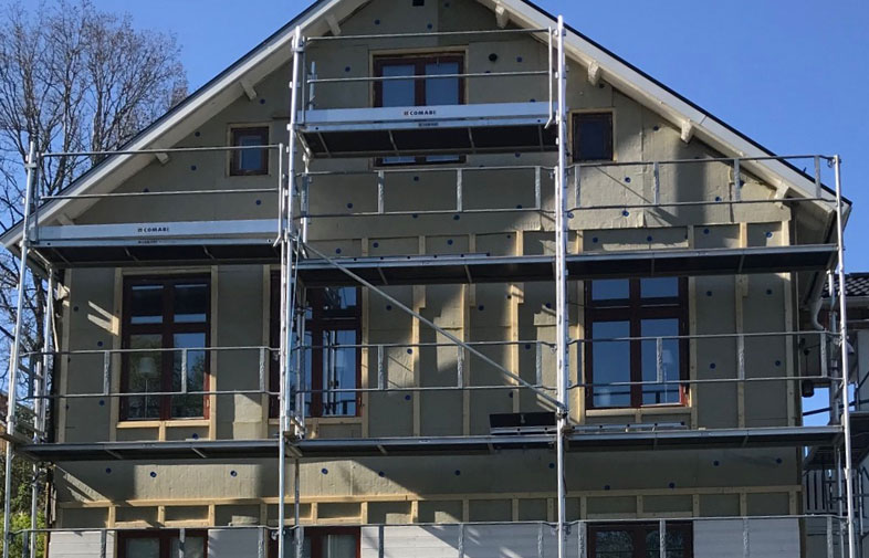 Beige flerfamiljshus med ställningar under renovering, mörkbruna fönster mot klarblå himmel