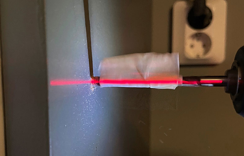 Rött laserljus träffar tejpad metallcylinder på grå vägg, dammpartiklar synliga.