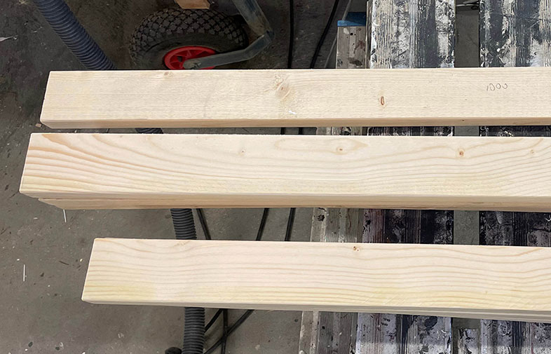 Tre ljusa träplankor på smutsig arbetsbänk i verkstad, rött hjul syns i bakgrunden