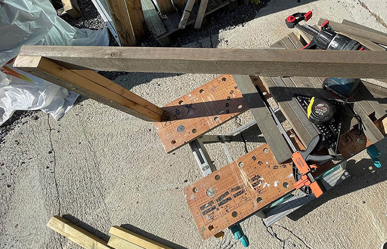 Utomhusarbetsbänk med bruna träplankor, orange borrmallar, svart-rött verktyg och måttband.