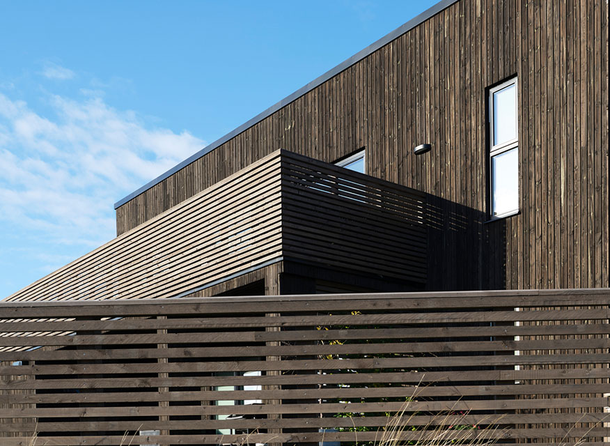 Modern mörk träfasad med horisontella ribbor, balkong och klarblå himmel.