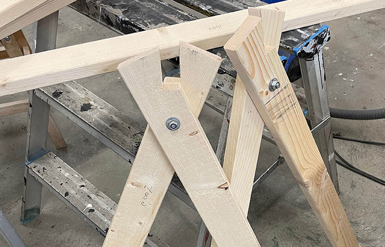 Två ljusa träbockar med metallbultar i en verkstad, svartmålad arbetsbock i bakgrunden.