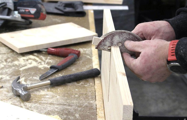 Händer slipar träplankans kant med halvmåne-slipdisk på dammig verkbänk med hammare och skruvmejsel.