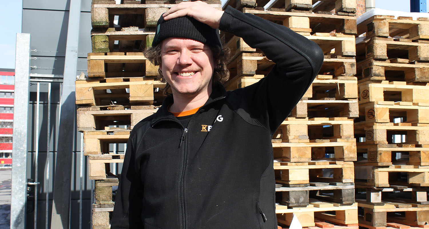 Person i svart jacka och mössa framför stapel av träpallar utomhus i industriell miljö.