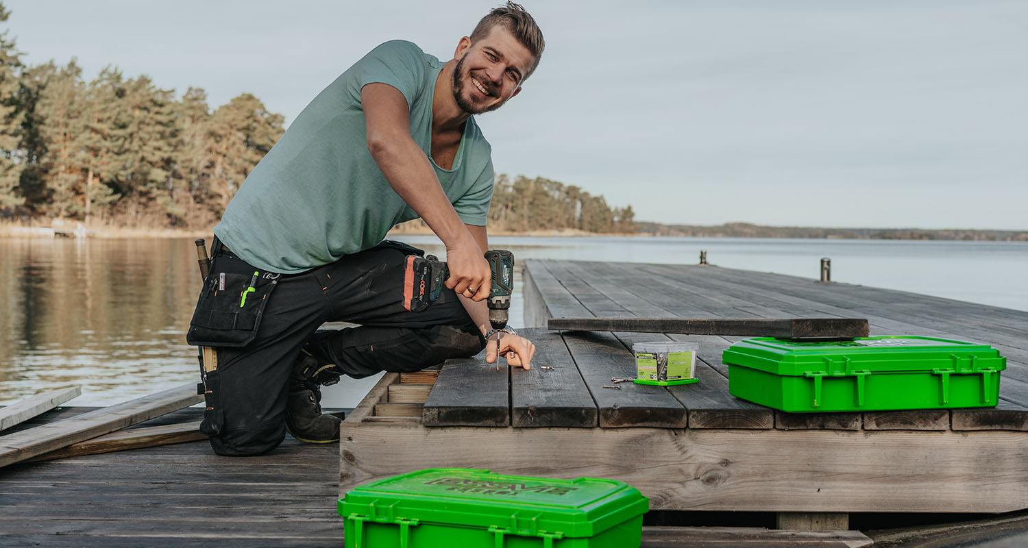 Man borrar trallbrädor på träbrygga vid stilla sjö, gröna verktygslådor bredvid.