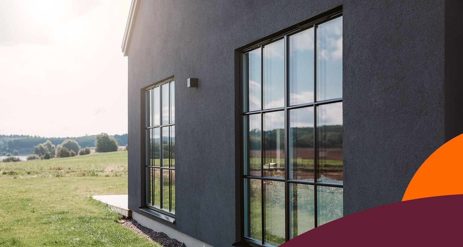 Modernt mörkgrått hus med stora rutindelade fönster som speglar grönt fält och blå himmel.