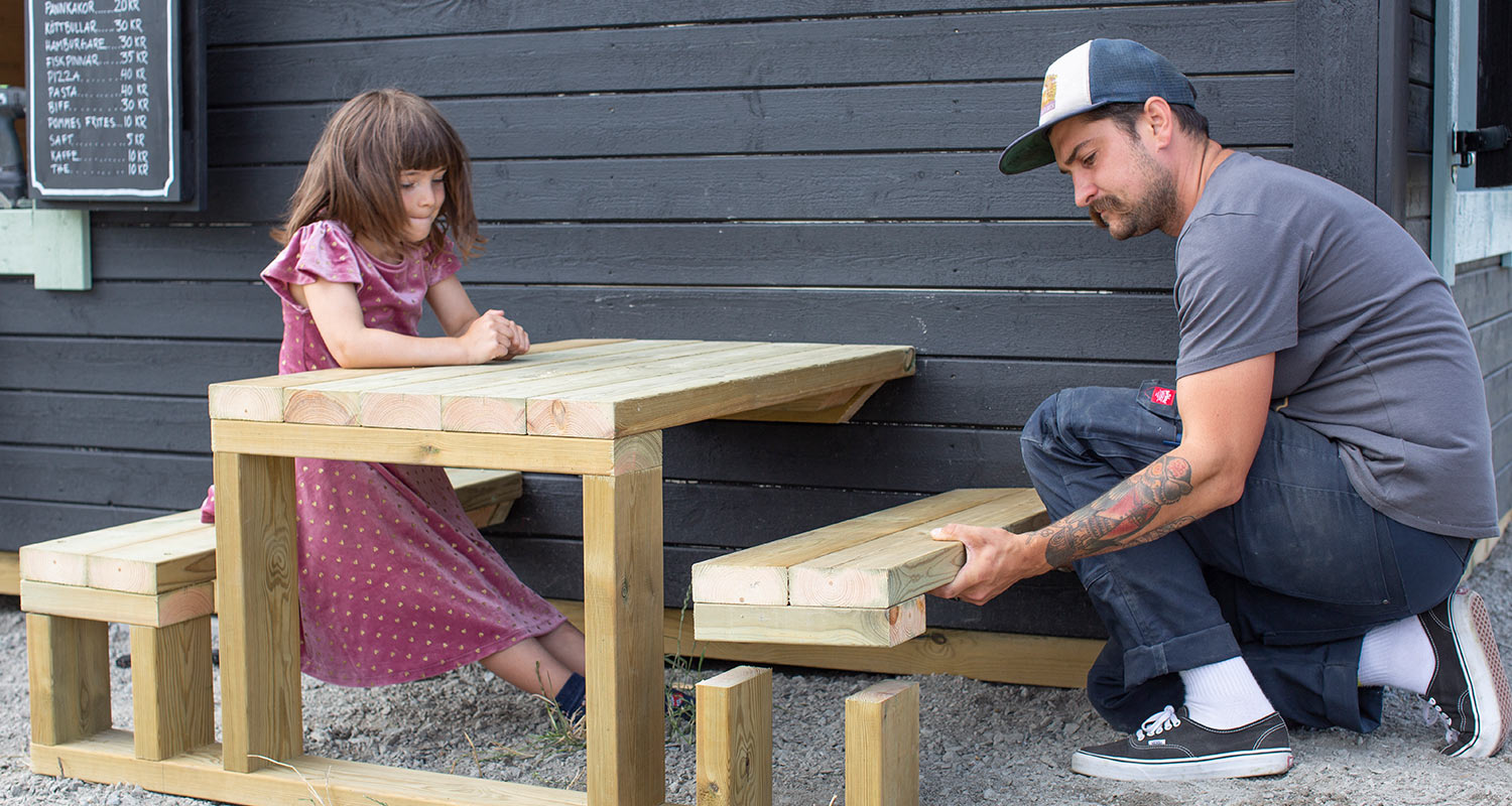 Man monterar ljus träbänk under bord vid svart träfasad, flicka i lila klänning väntar.