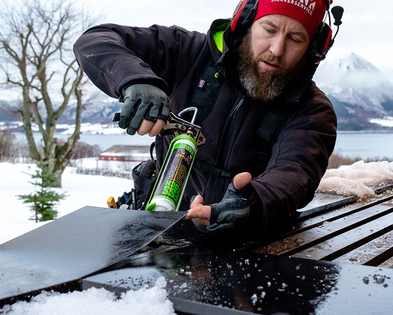 Arbetare i röd hjälm applicerar tätningsmassa på svart plåt i snölandskap vid fjord
